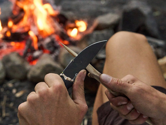 Hands carving a wooden stick with a black bladed CIVIVI knife beside a campfire outdoors