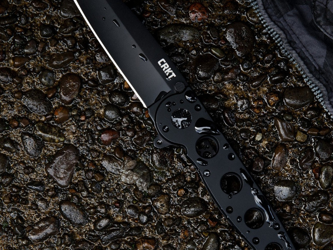 CRKT folding knife with a black coated tanto blade lying on wet river stones beside a jacket