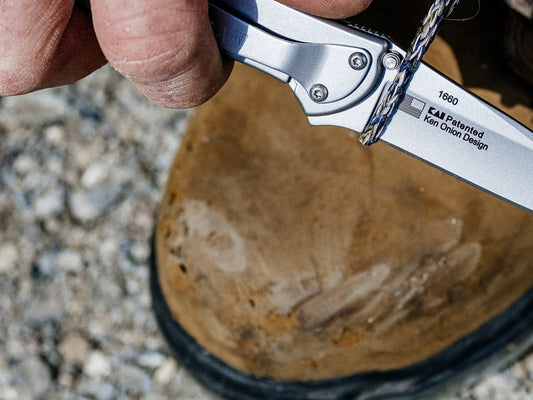 Kershaw Leek folding knife held open in one hand above a worn leather boot on gravel