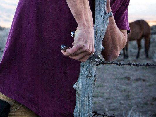Man using a SOG multi-tool to twist barbed wire on a wooden fence post with horses grazing in the background
