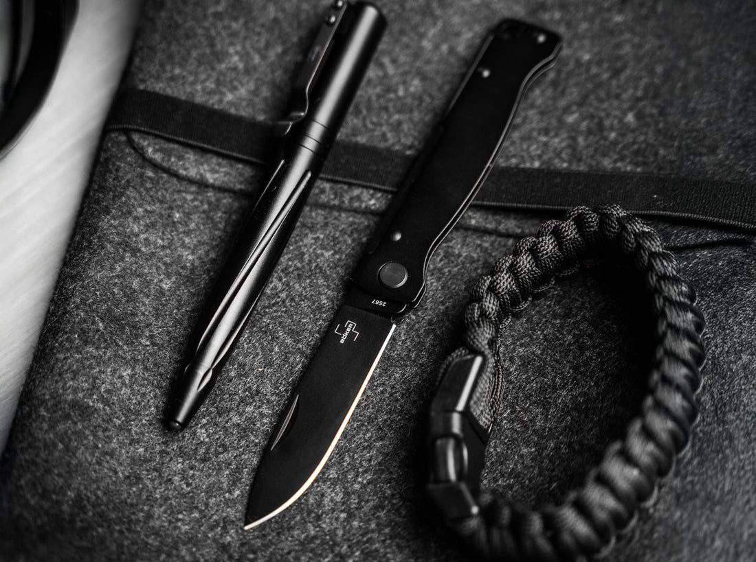 Everyday carry flat lay with a black slipjoint pocket knife, a black tactical pen and a paracord bracelet on grey felt