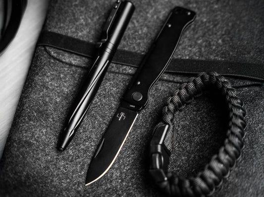 Everyday carry flat lay with a black slipjoint pocket knife, a black tactical pen and a paracord bracelet on grey felt