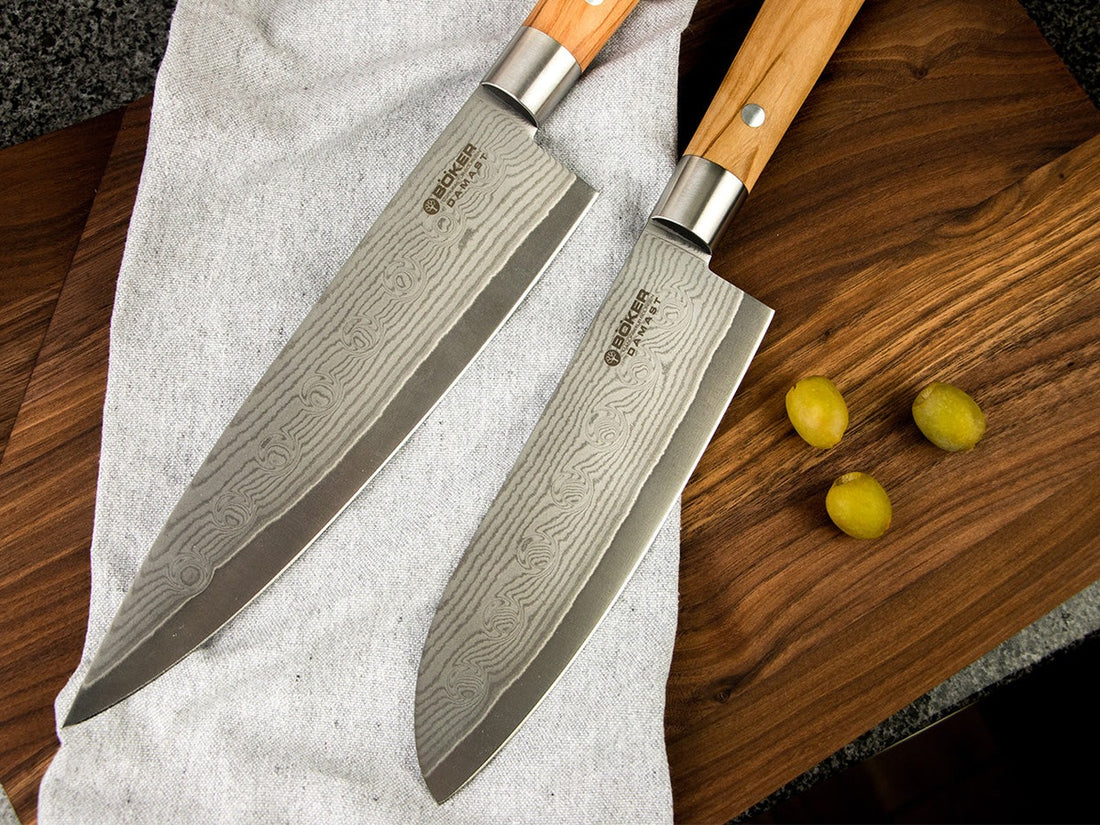 Two Damascus chef's knives with olive wood handles resting on a linen cloth and a walnut cutting board in a kitchen