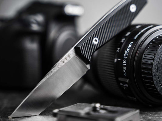 Tanto-point EDC knife blade resting on a camera lens on a dark leather desk mat